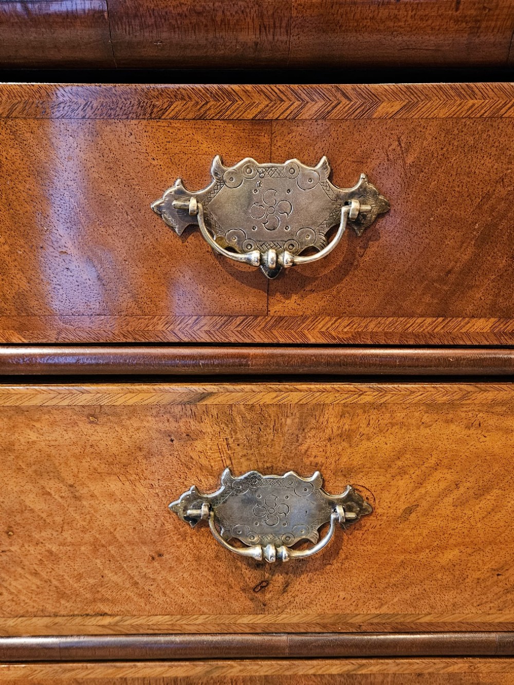 A George I walnut chest of drawers featuring herringbone crossbanding, brass engraved hardware, and bun feet against a dark wall. View V