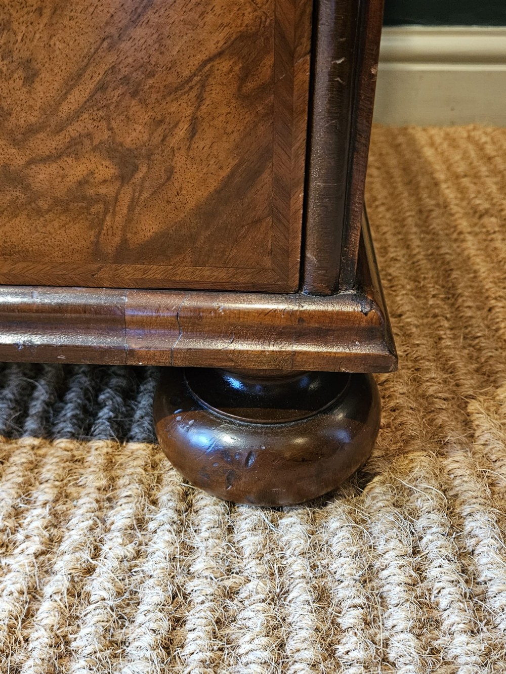 A George I walnut chest of drawers featuring herringbone crossbanding, brass engraved hardware, and bun feet against a dark wall. View VI