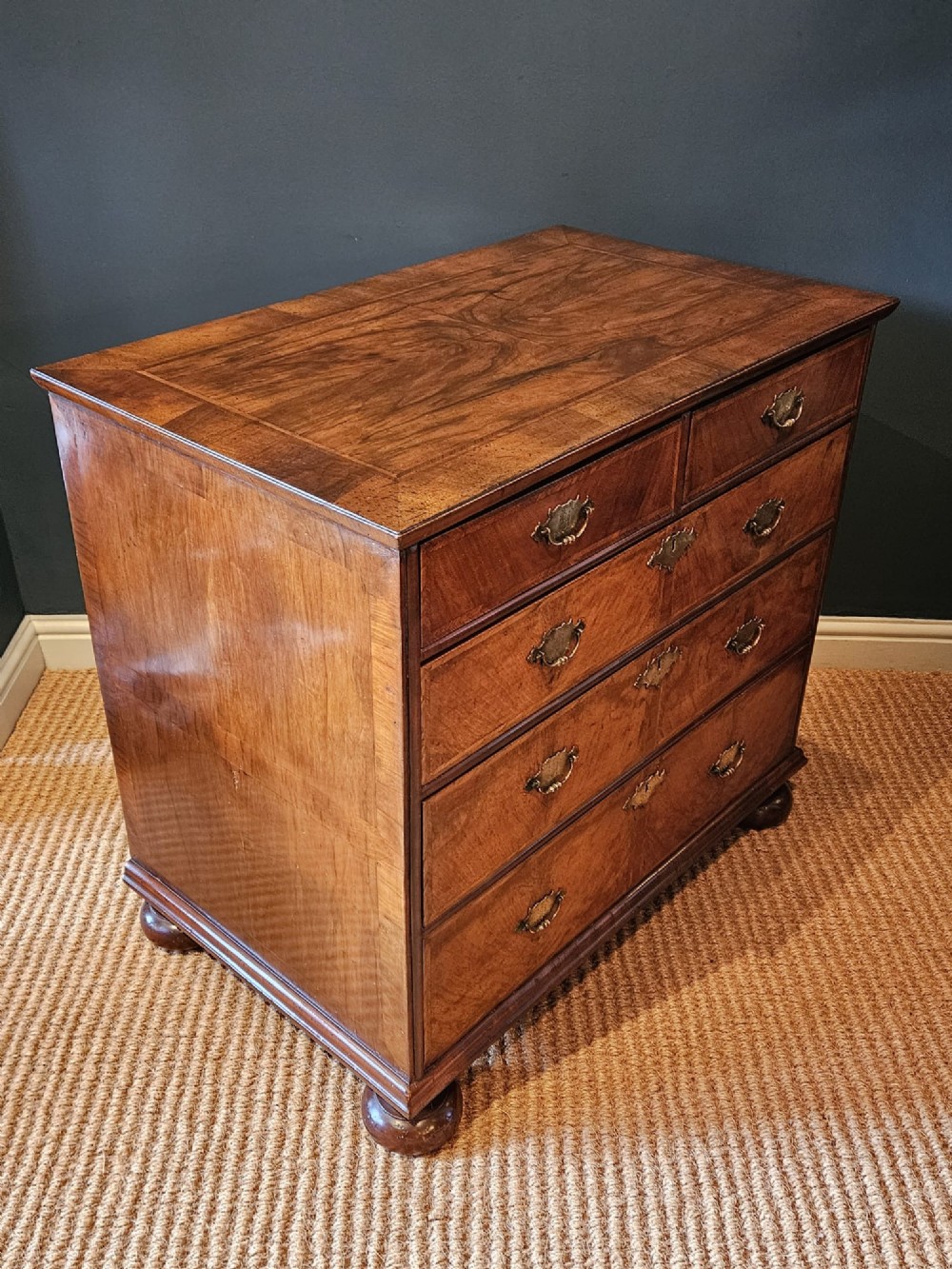 A George I walnut chest of drawers featuring herringbone crossbanding, brass engraved hardware, and bun feet against a dark wall. View VII