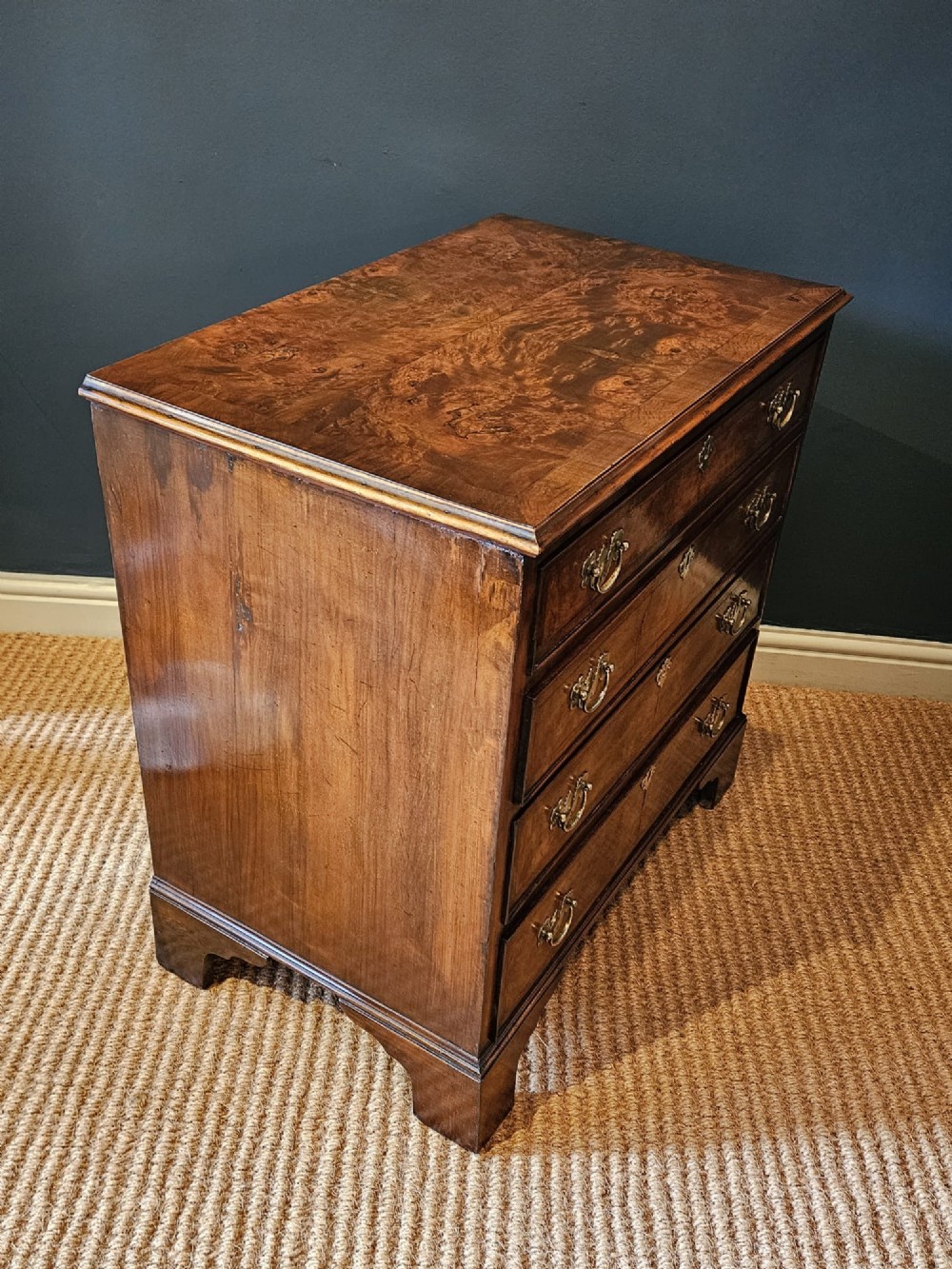 A small George III walnut chest of drawers featuring four graduated drawers with brass handles, keyhole escutcheons, and bracket feet against a dark grey background. View III