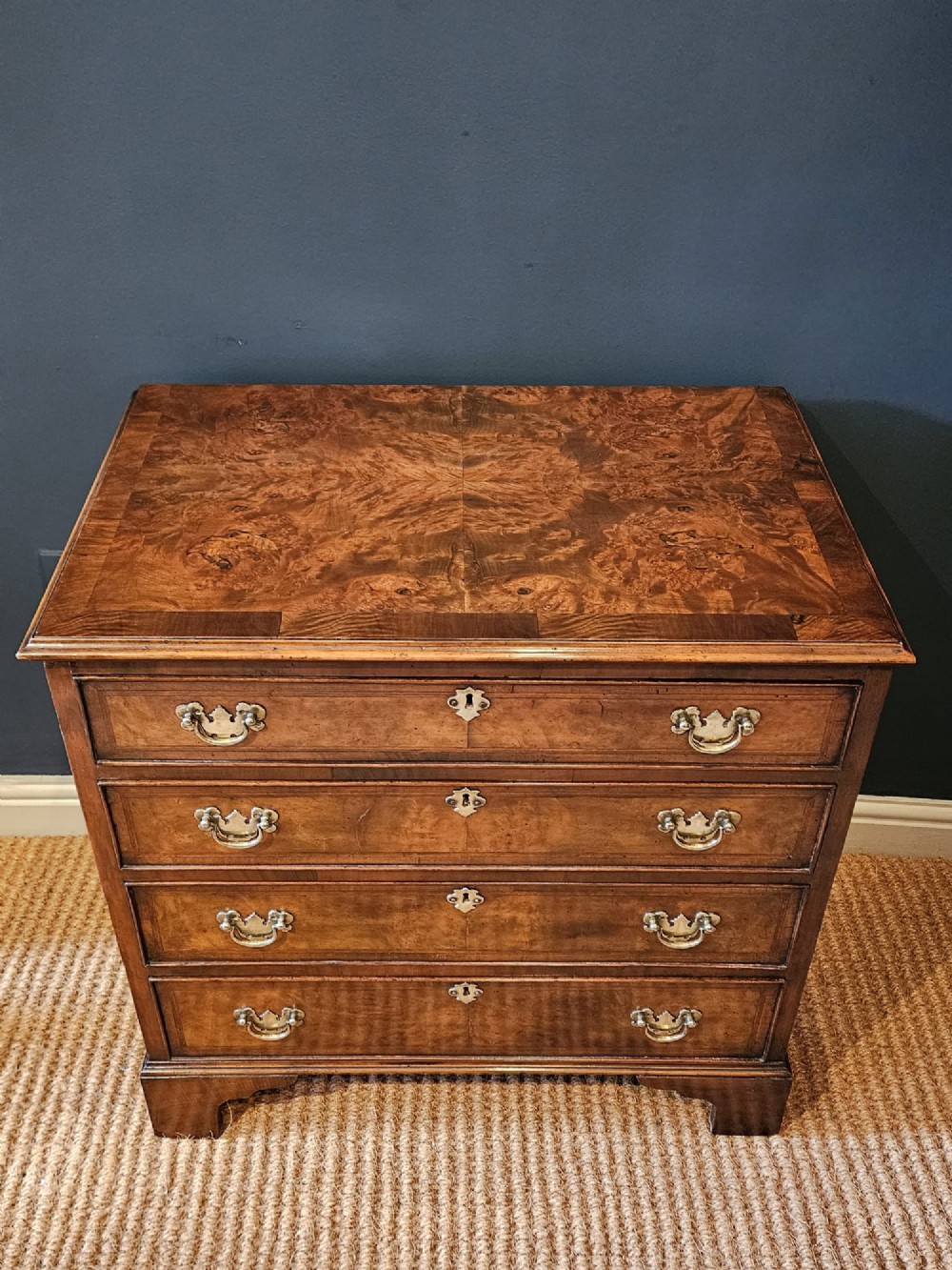 A small George III walnut chest of drawers featuring four graduated drawers with brass handles, keyhole escutcheons, and bracket feet against a dark grey background. View IV