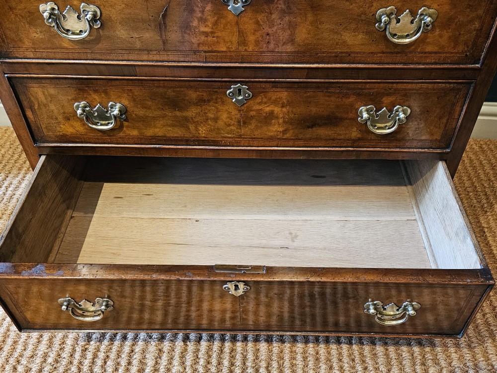 A small George III walnut chest of drawers featuring four graduated drawers with brass handles, keyhole escutcheons, and bracket feet against a dark grey background. View V