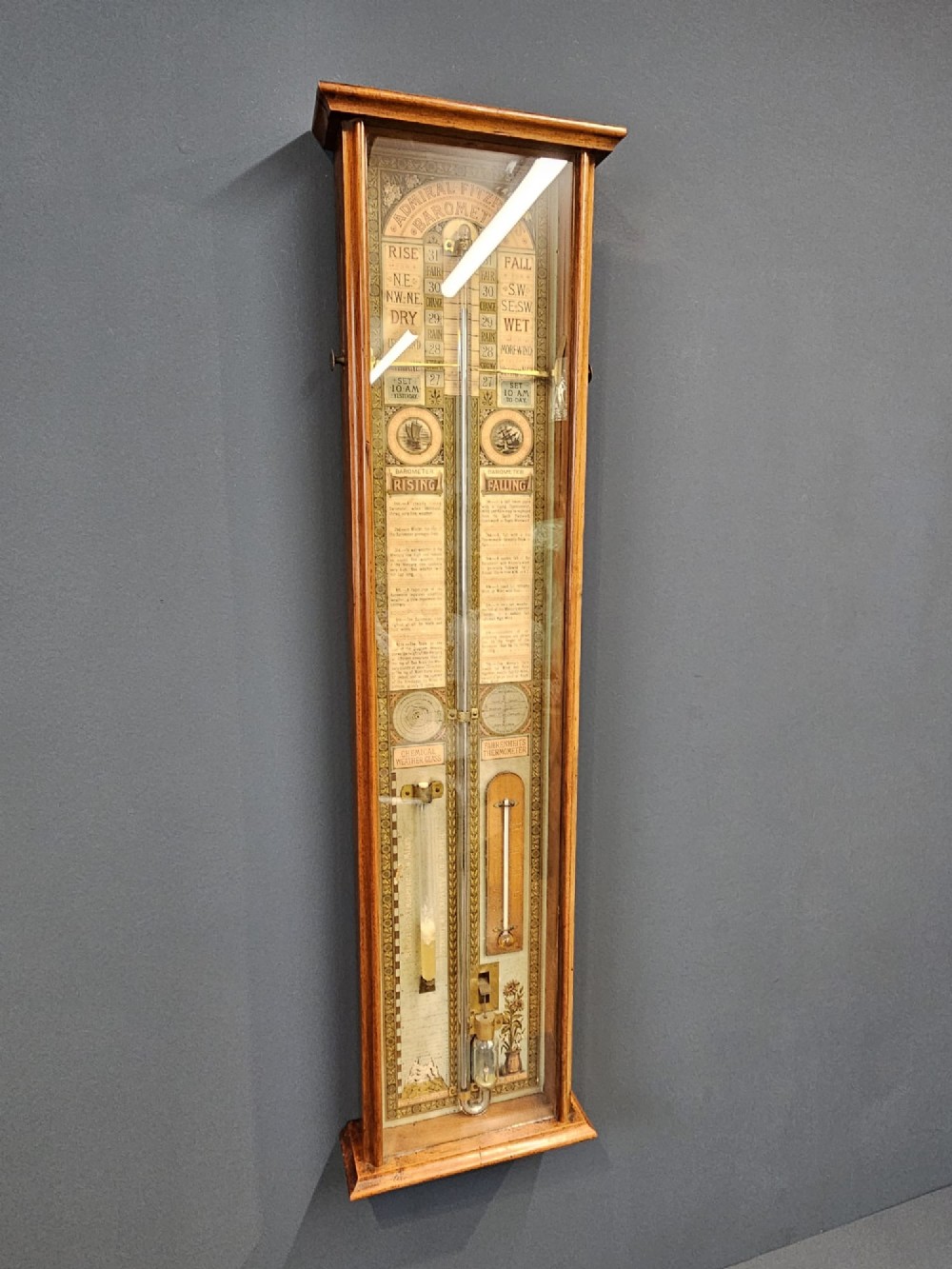 A glazed timber-cased Victorian Admiral Fitzroy wall barometer featuring printed weather prediction scales, a central glass barometer tube, a thermometer, and a storm glass. View V