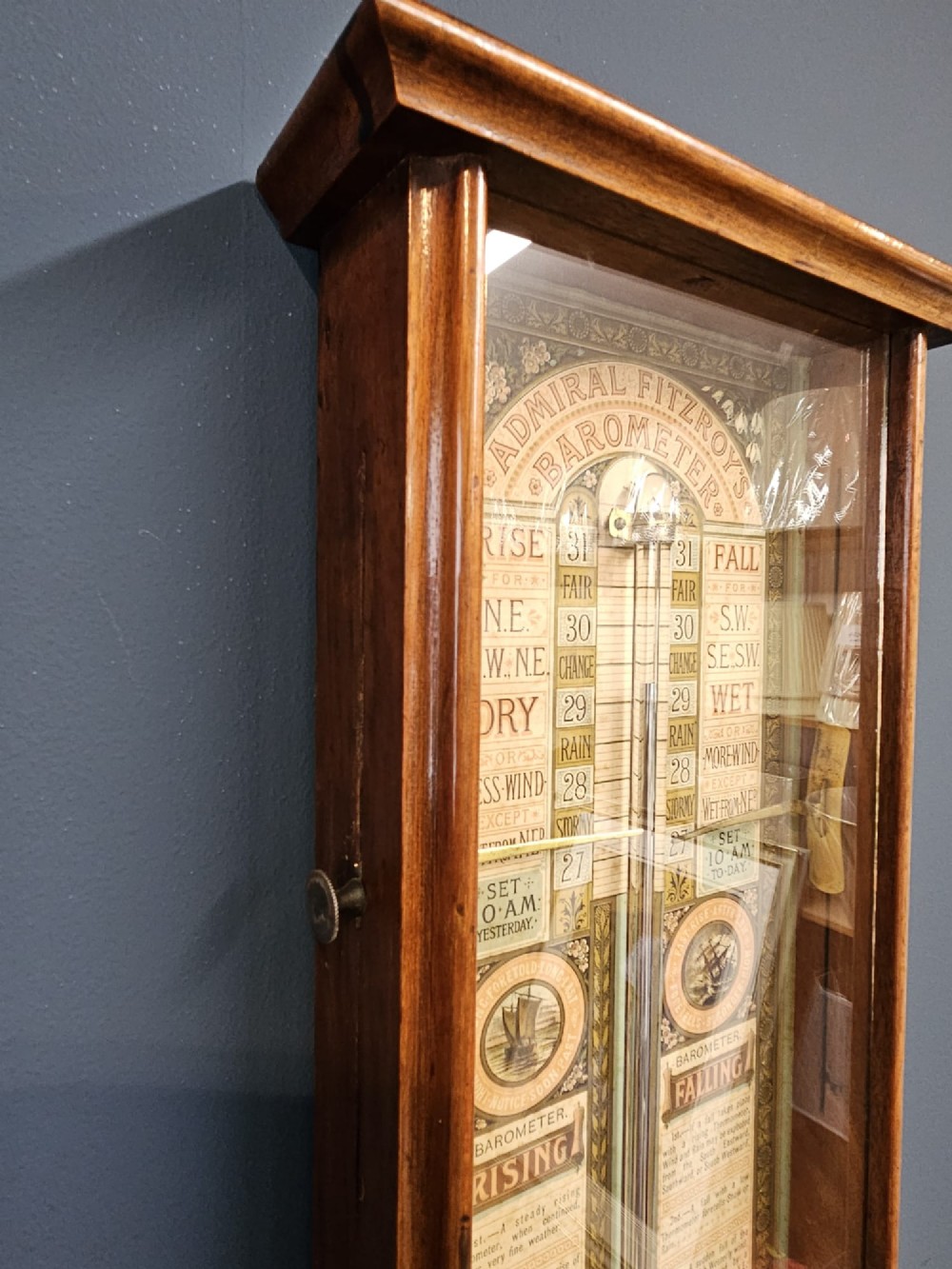 A glazed timber-cased Victorian Admiral Fitzroy wall barometer featuring printed weather prediction scales, a central glass barometer tube, a thermometer, and a storm glass. View VI