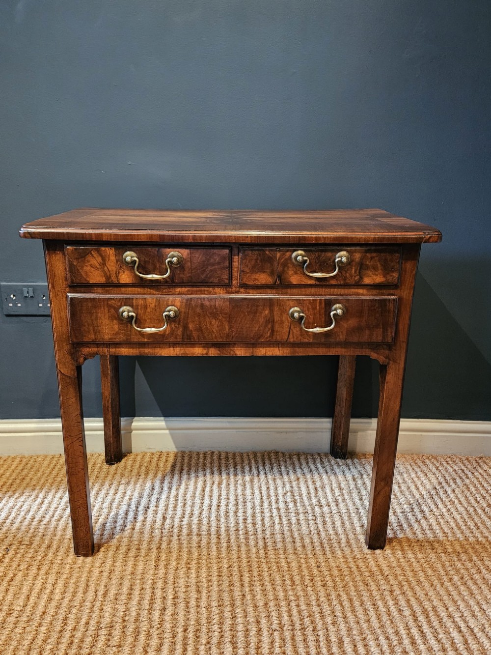 A Georgian walnut side table featuring three drawers with brass handles and straight legs set against a dark blue wall.