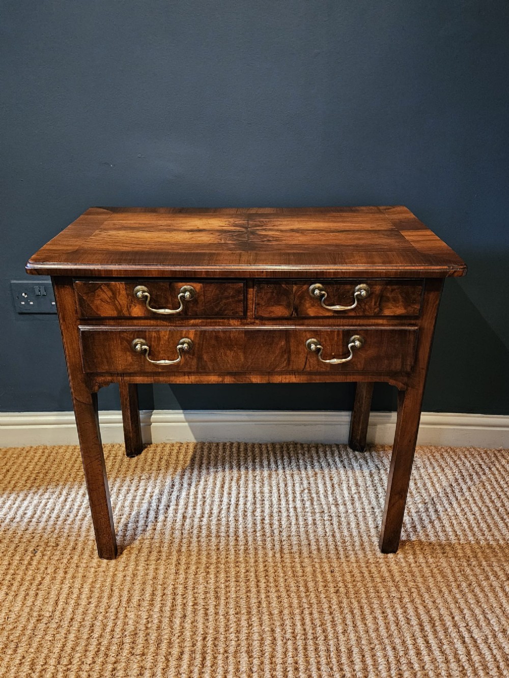 A Georgian walnut side table featuring three drawers with brass handles and straight legs set against a dark blue wall. View II
