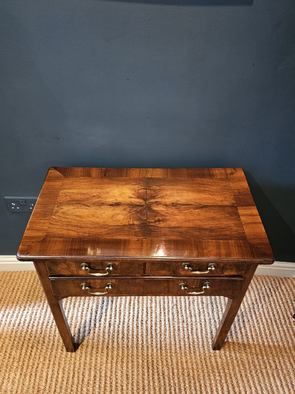 A Georgian walnut side table featuring three drawers with brass handles and straight legs set against a dark blue wall. View IV