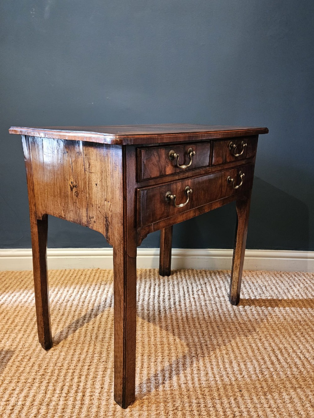 A Georgian walnut side table featuring three drawers with brass handles and straight legs set against a dark blue wall. View VI