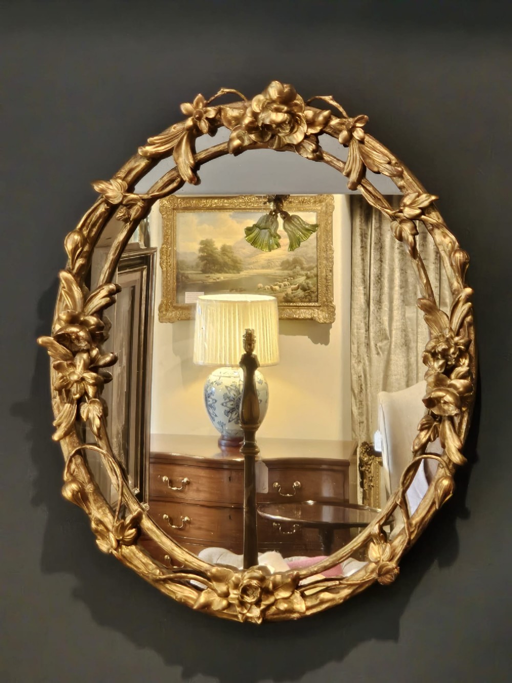 An oval Victorian giltwood wall mirror featuring a frame intricately carved with rose blossoms, foliage, and vine motifs. View VI