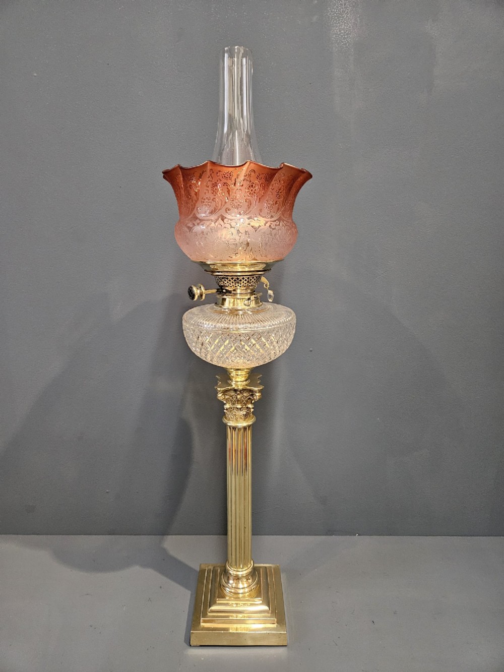 A Victorian oil lamp featuring a stepped brass base, fluted Corinthian column stem, cut crystal reservoir, etched pink glass shade, and clear glass chimney.