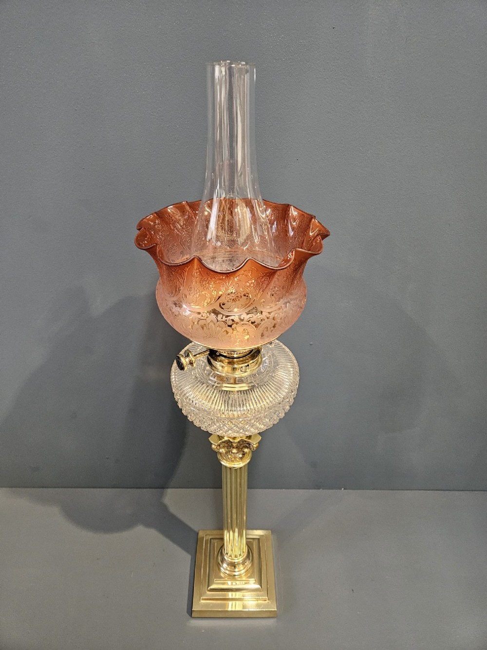 A Victorian oil lamp featuring a stepped brass base, fluted Corinthian column stem, cut crystal reservoir, etched pink glass shade, and clear glass chimney. View VI