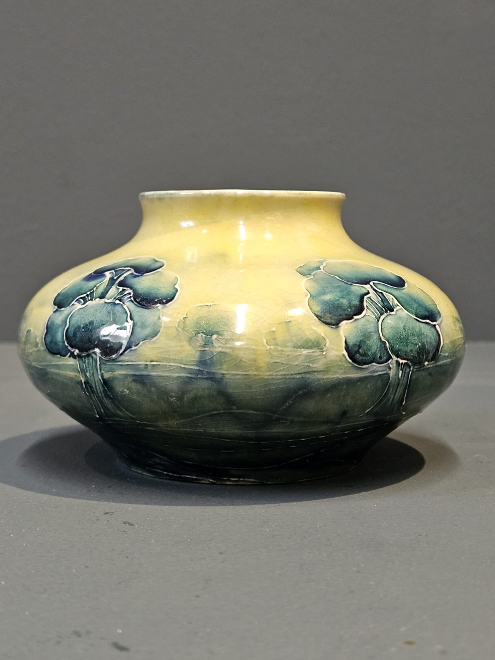 A squat ceramic Art Nouveau vase featuring a hand-painted tube-lined tree motif in shades of teal blue and green over a soft yellow background. View II