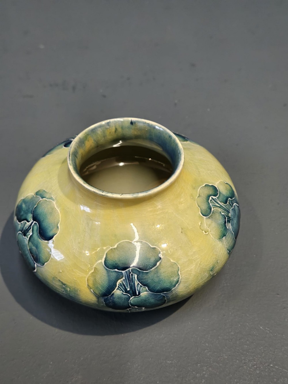 A squat ceramic Art Nouveau vase featuring a hand-painted tube-lined tree motif in shades of teal blue and green over a soft yellow background. View VI