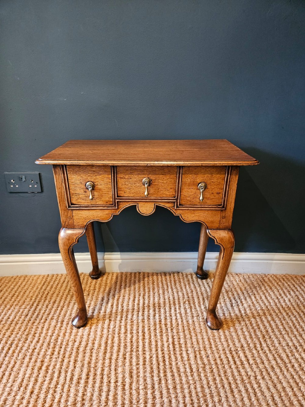 A small George II oak lowboy featuring three drawers with brass teardrop handles, a shaped apron, and cabriole legs terminating in pad feet.