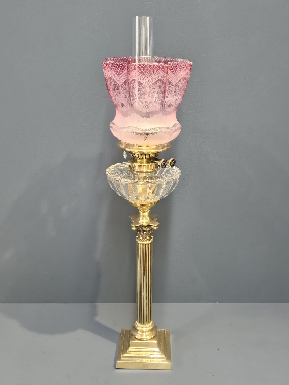 A Victorian brass oil lamp featuring a stepped square base, fluted Corinthian column, cut glass reservoir, and a flared etched cranberry glass shade.