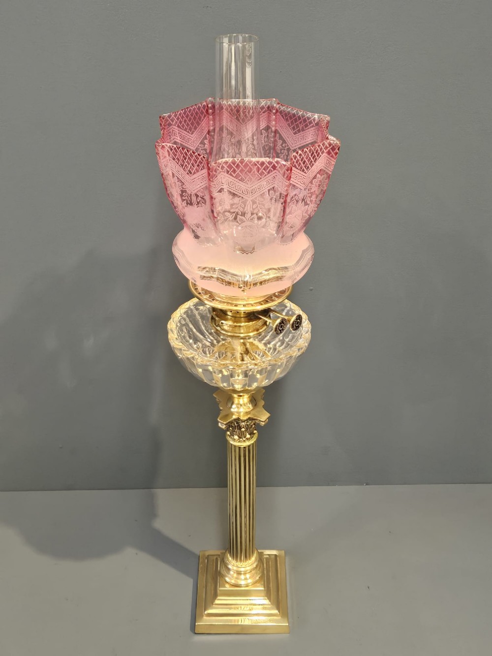 A Victorian brass oil lamp featuring a stepped square base, fluted Corinthian column, cut glass reservoir, and a flared etched cranberry glass shade. View VI