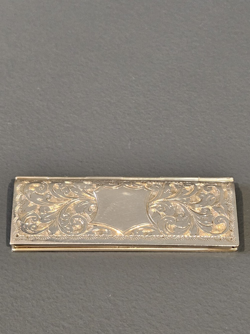 A slim rectangular silver box featuring intricate engraved foliate scrollwork and a central unengraved shield cartouche.