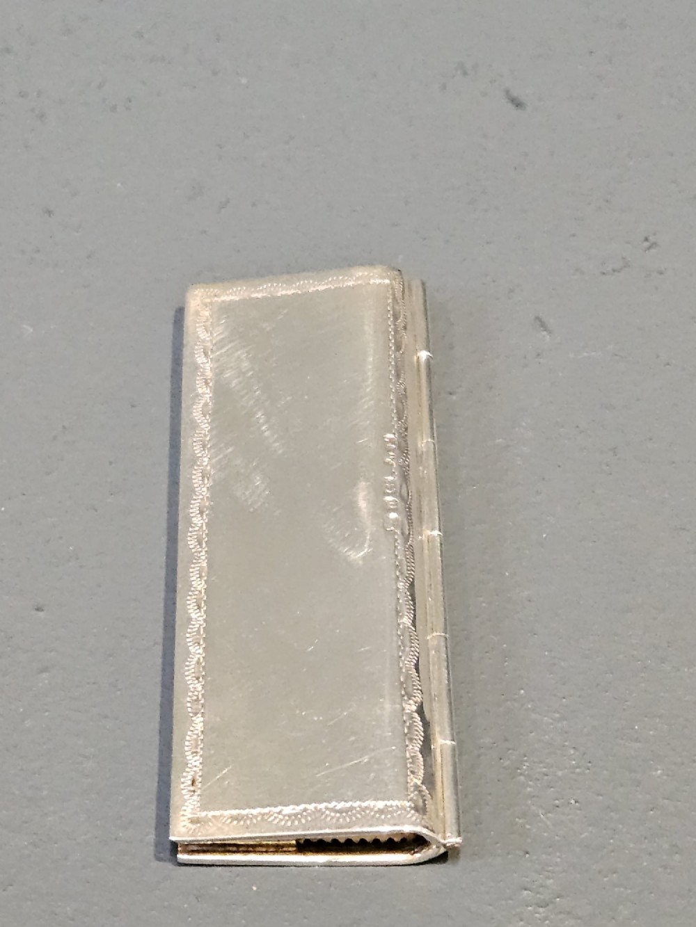 A slim rectangular silver box featuring intricate engraved foliate scrollwork and a central unengraved shield cartouche. View II