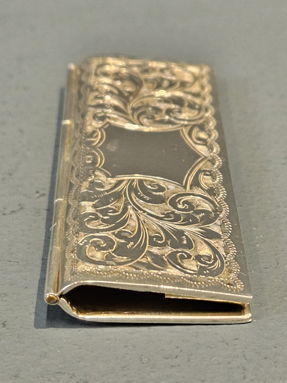 A slim rectangular silver box featuring intricate engraved foliate scrollwork and a central unengraved shield cartouche. View III