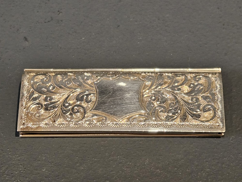 A slim rectangular silver box featuring intricate engraved foliate scrollwork and a central unengraved shield cartouche. View VI