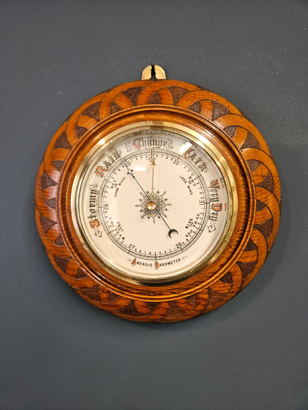 A circular Victorian aneroid wall barometer housed in a turned and carved oak case with a brass bezel. View II