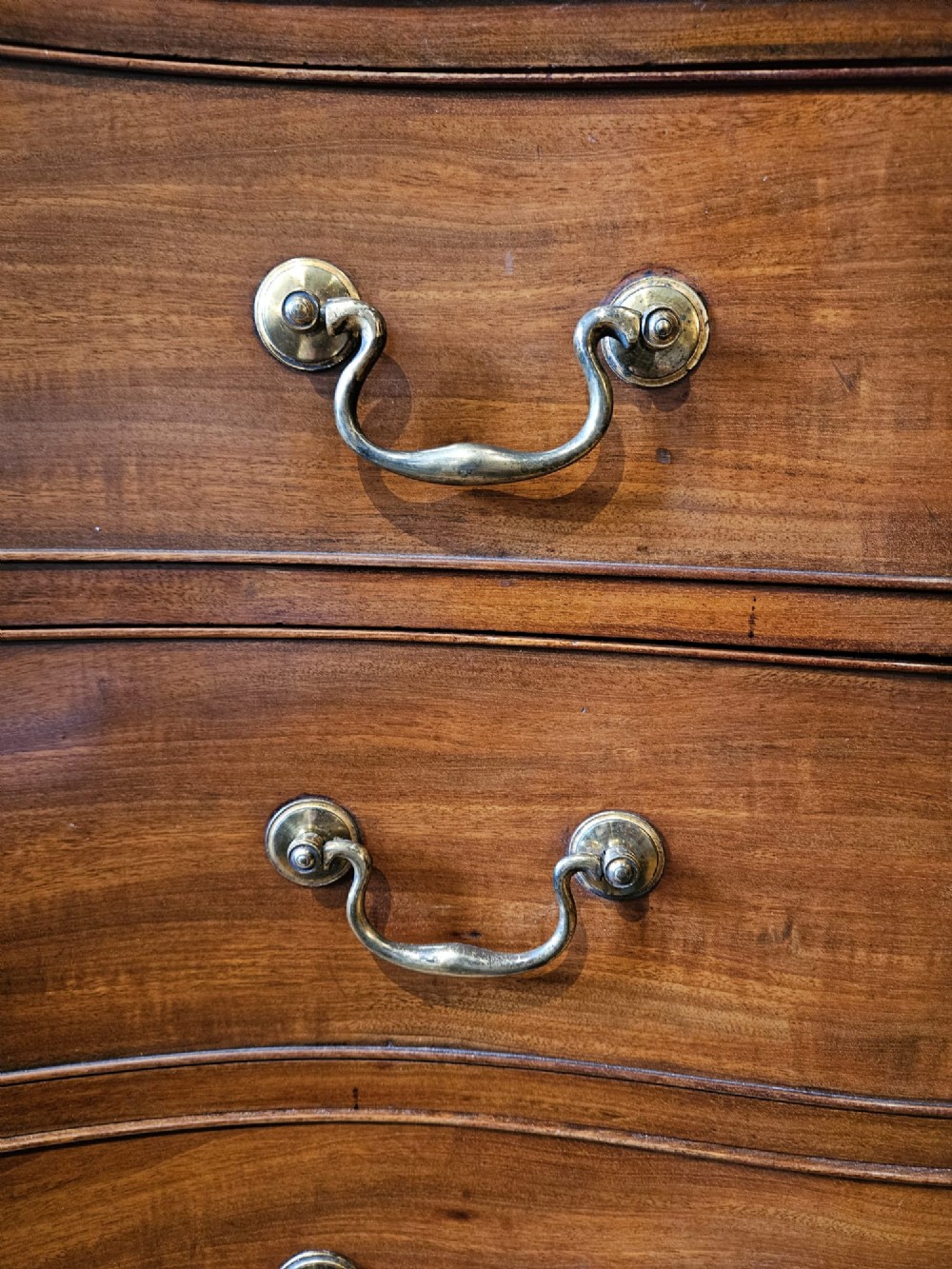 A George III mahogany chest of drawers featuring a curved serpentine front, four graduated drawers with brass swan-neck handles, and splayed feet. View II