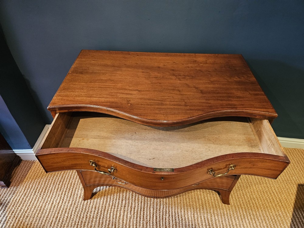 A George III mahogany chest of drawers featuring a curved serpentine front, four graduated drawers with brass swan-neck handles, and splayed feet. View VII