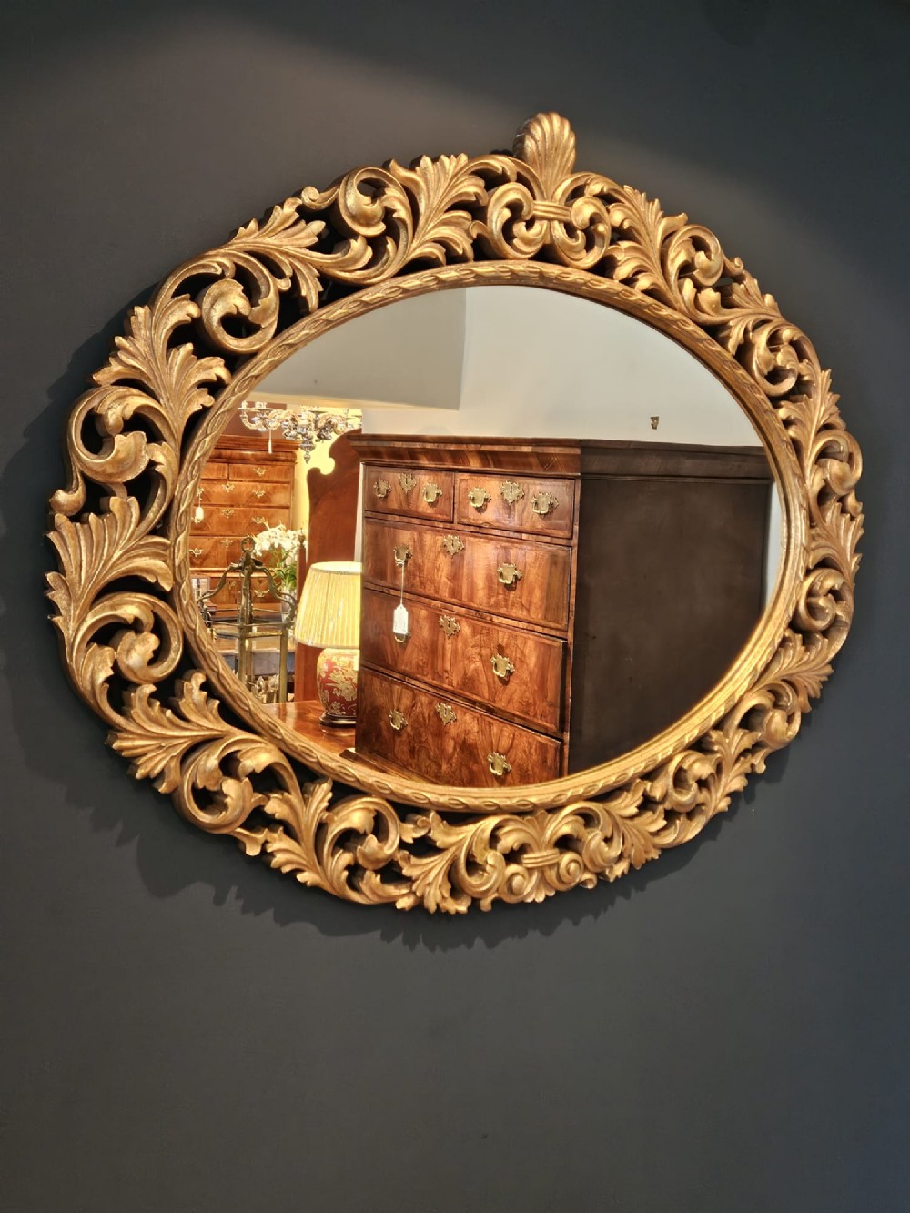 An ornate oval wall mirror featuring a heavily carved timber frame with gold gilt finish, decorated with openwork acanthus leaf scrolls and a top cresting.