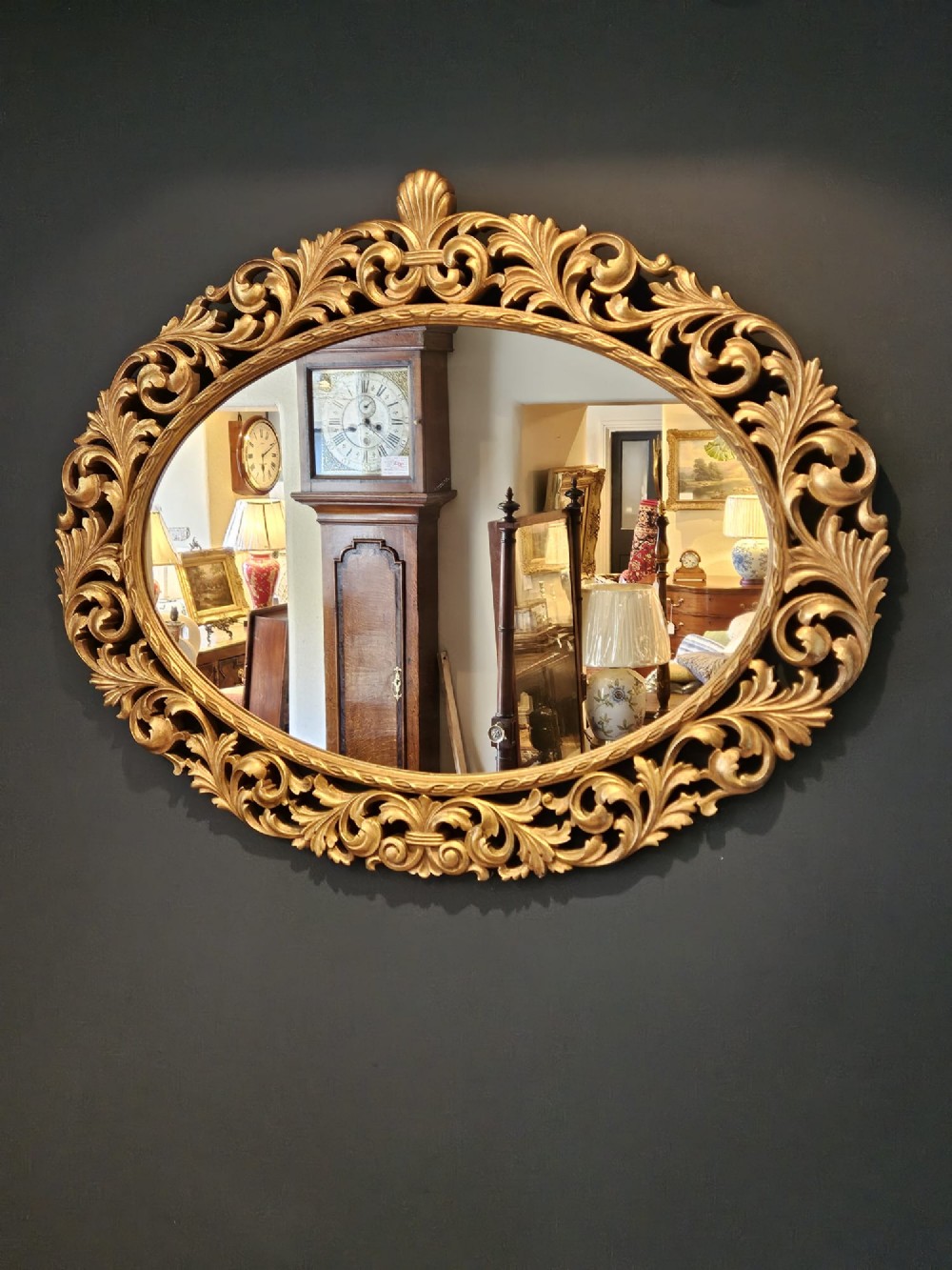 An ornate oval wall mirror featuring a heavily carved timber frame with gold gilt finish, decorated with openwork acanthus leaf scrolls and a top cresting. View II