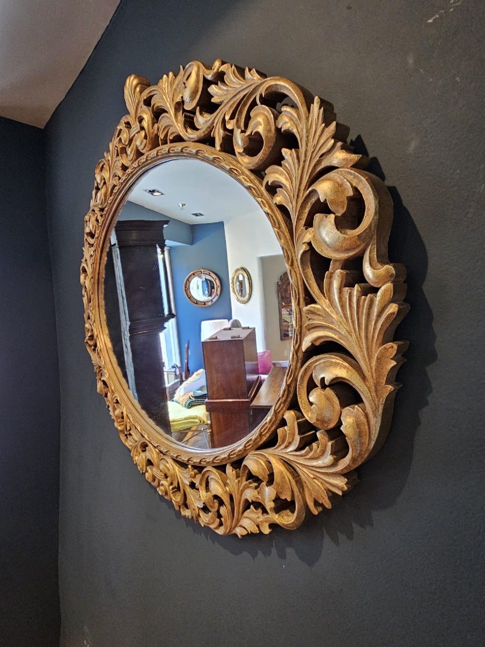 An ornate oval wall mirror featuring a heavily carved timber frame with gold gilt finish, decorated with openwork acanthus leaf scrolls and a top cresting. View IV