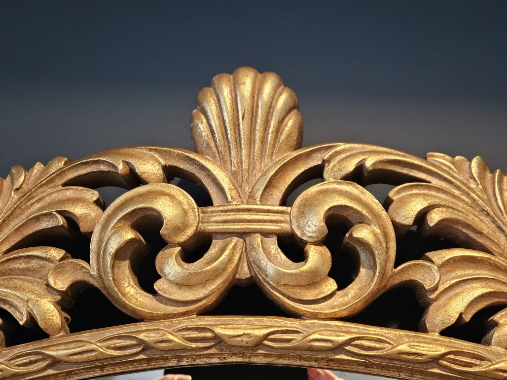 An ornate oval wall mirror featuring a heavily carved timber frame with gold gilt finish, decorated with openwork acanthus leaf scrolls and a top cresting. View V