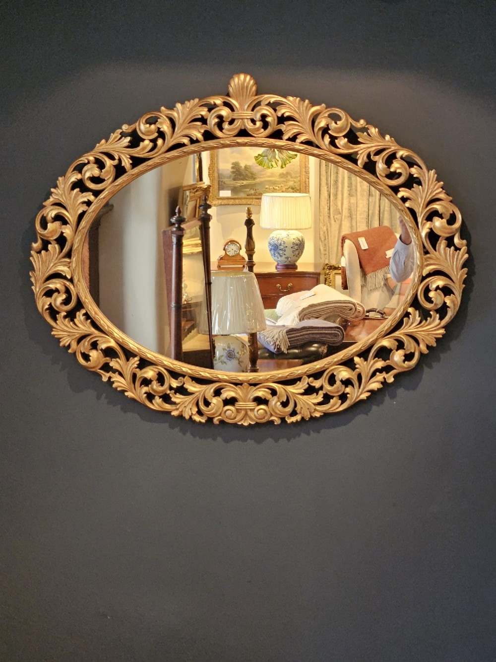 An ornate oval wall mirror featuring a heavily carved timber frame with gold gilt finish, decorated with openwork acanthus leaf scrolls and a top cresting. View VI