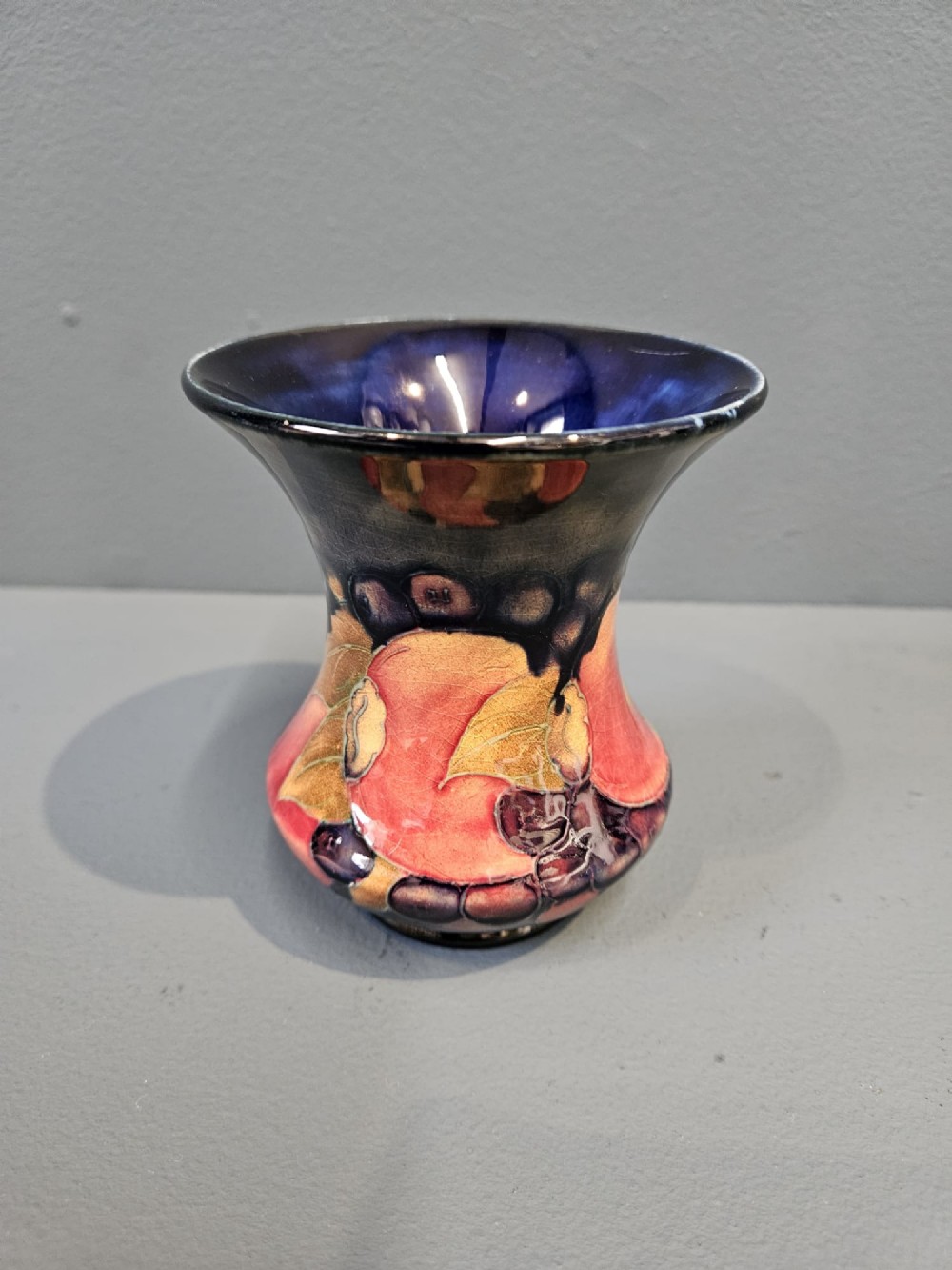 An Art Nouveau Moorcroft earthenware vase with a flared rim, featuring hand-painted tube-lined pomegranates and berries against a deep cobalt blue background.