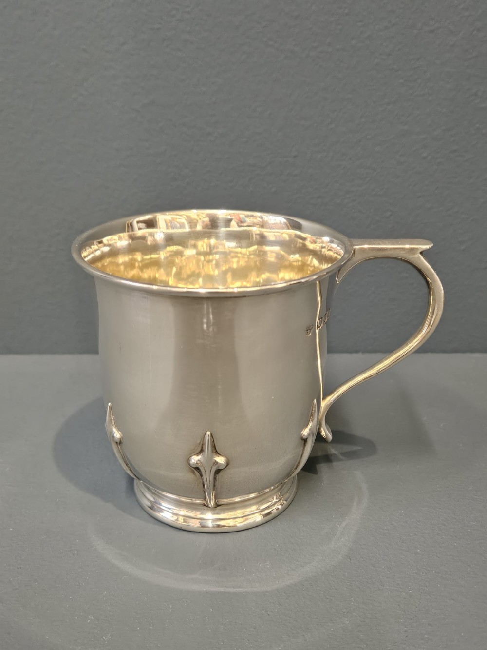 A hallmarked sterling silver mug with raised strapwork details around the lower body, a gilded interior, and a single loop handle.