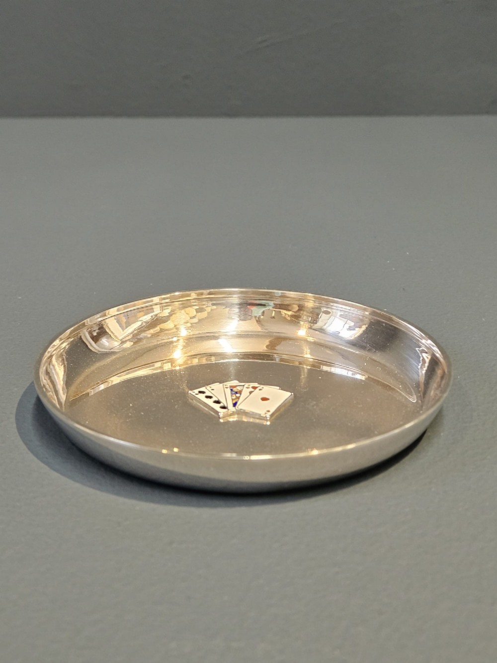 A circular sterling silver dish featuring a central polychrome enamel playing card fan design and stamped British hallmarks. View IV