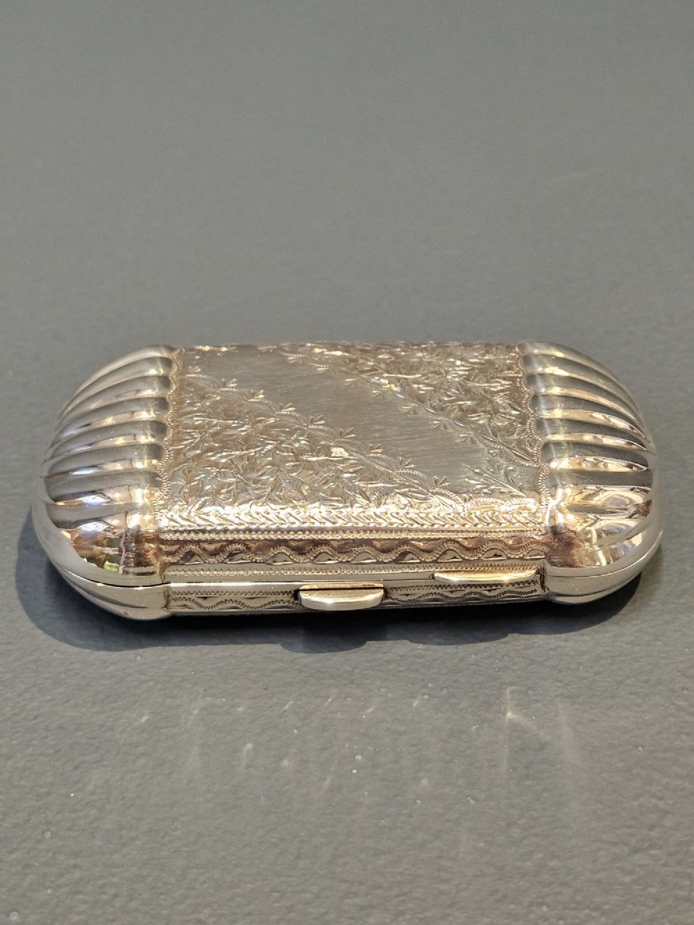 A small rectangular Victorian sterling silver cheroot case featuring hand-engraved foliate decoration and ribbed fluted ends on a grey surface.