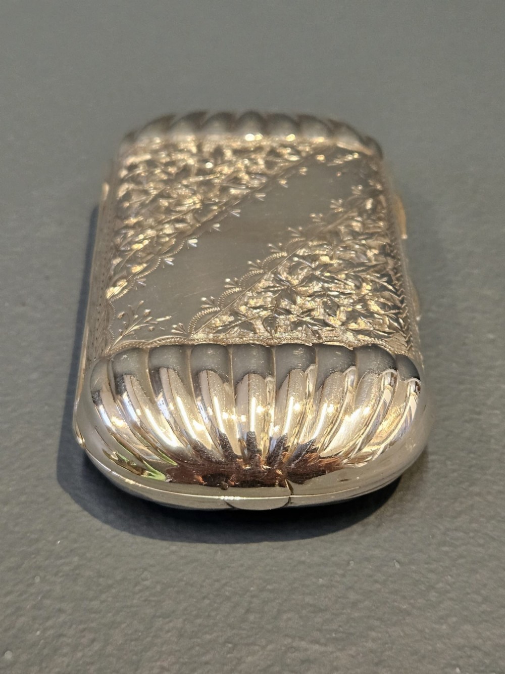 A small rectangular Victorian sterling silver cheroot case featuring hand-engraved foliate decoration and ribbed fluted ends on a grey surface. View II