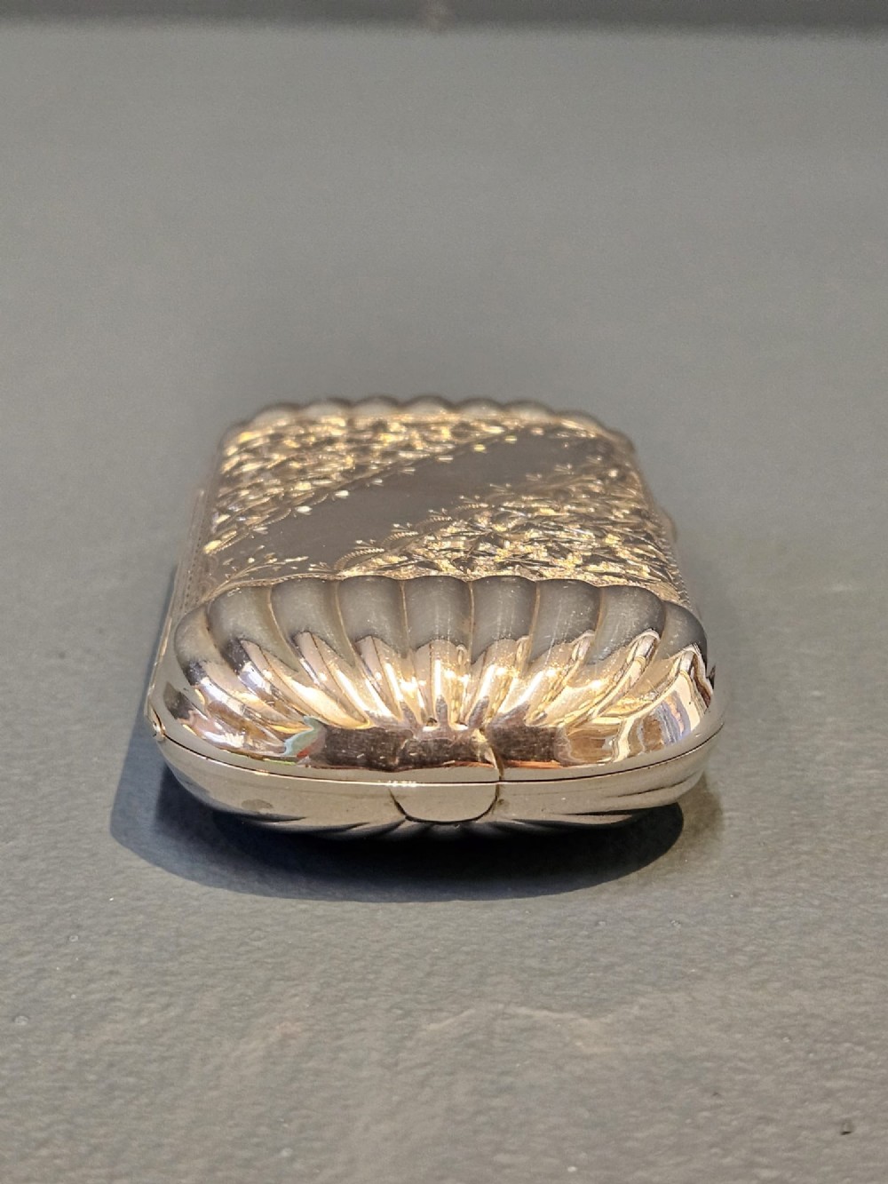A small rectangular Victorian sterling silver cheroot case featuring hand-engraved foliate decoration and ribbed fluted ends on a grey surface. View V