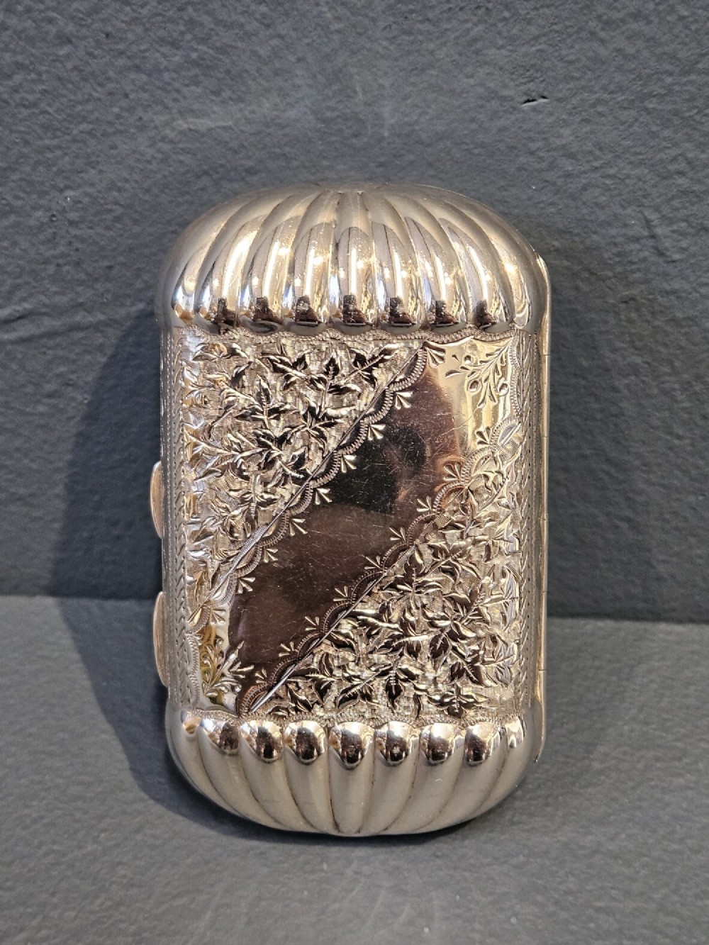A small rectangular Victorian sterling silver cheroot case featuring hand-engraved foliate decoration and ribbed fluted ends on a grey surface. View VII