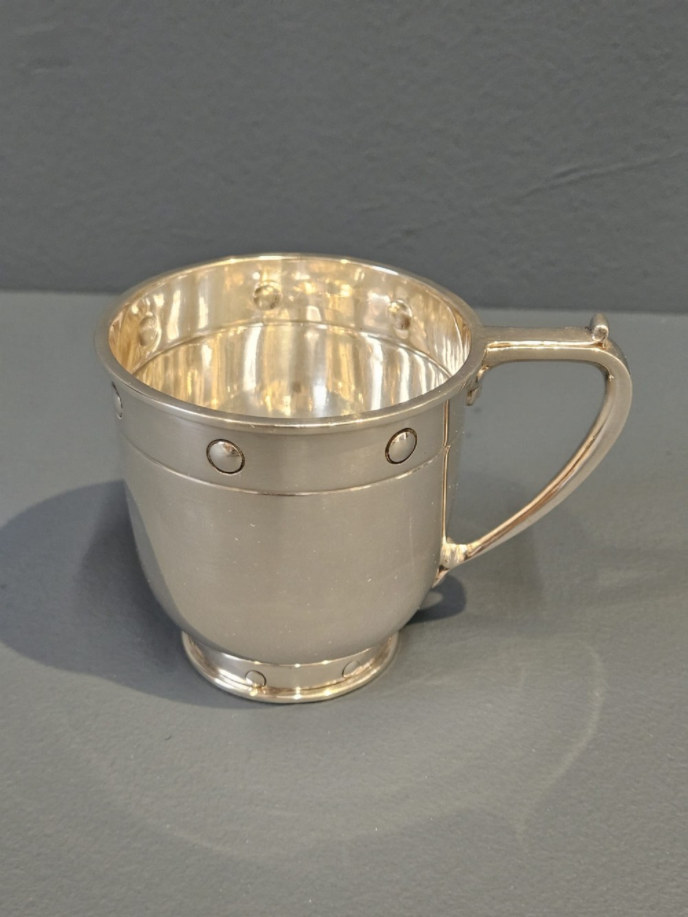 A polished sterling silver George V mug featuring riveted border detailing and a shaped handle. View II
