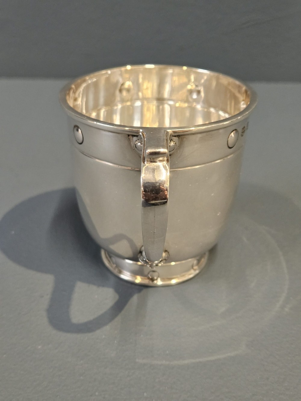 A polished sterling silver George V mug featuring riveted border detailing and a shaped handle. View III
