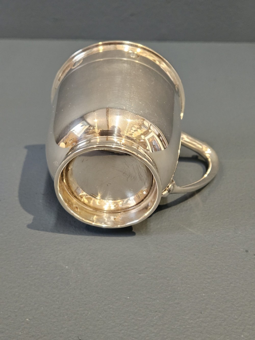 A polished sterling silver George V mug featuring riveted border detailing and a shaped handle. View IV