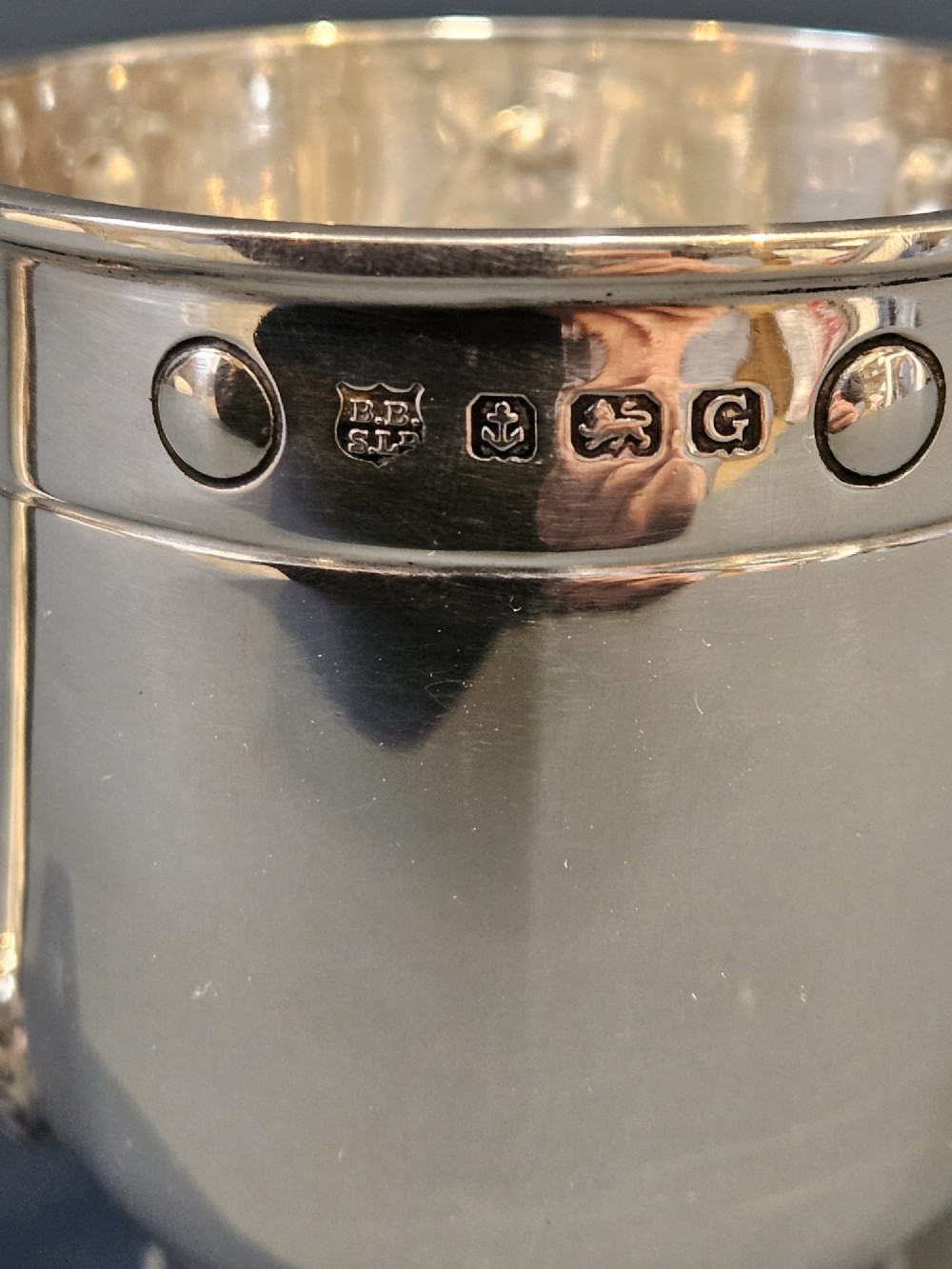 A polished sterling silver George V mug featuring riveted border detailing and a shaped handle. View V