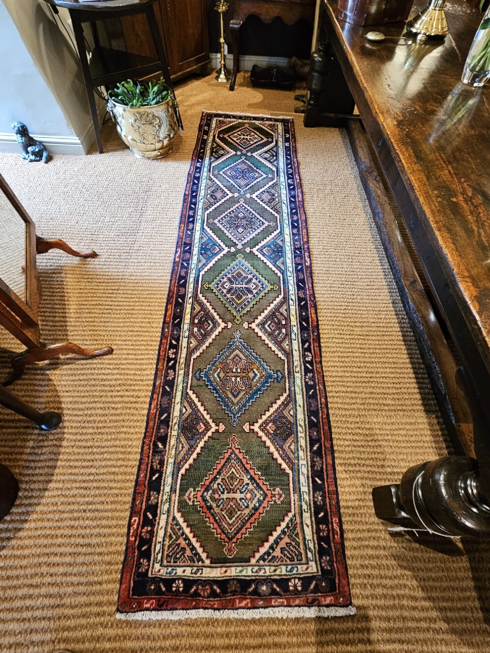 A long hand-woven wool runner rug featuring repeating diamond geometric medallions on an olive green background with decorative navy borders.