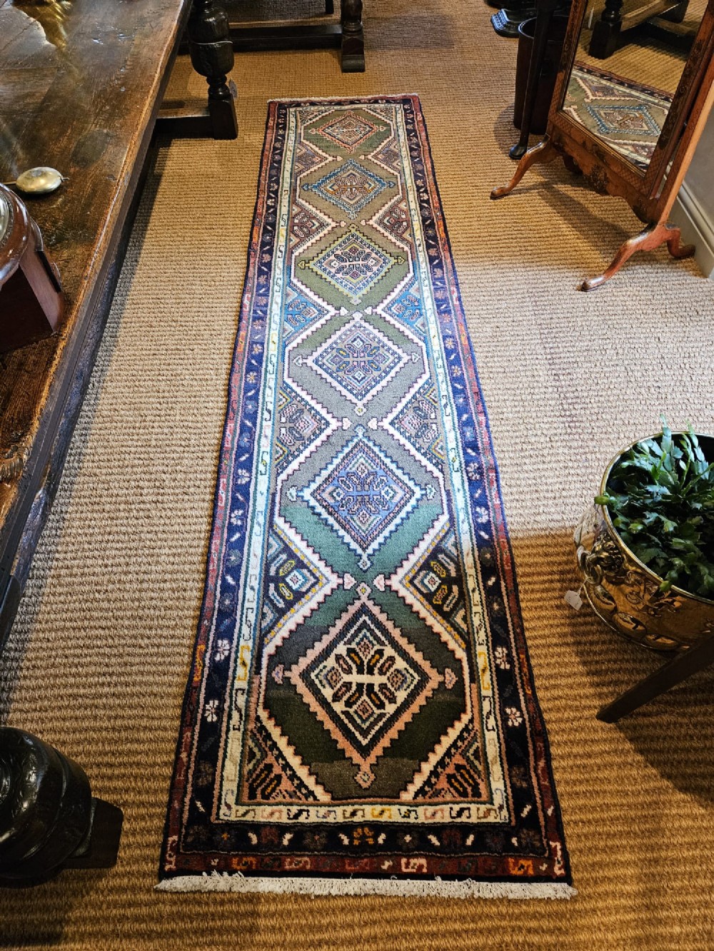 A long hand-woven wool runner rug featuring repeating diamond geometric medallions on an olive green background with decorative navy borders. View II