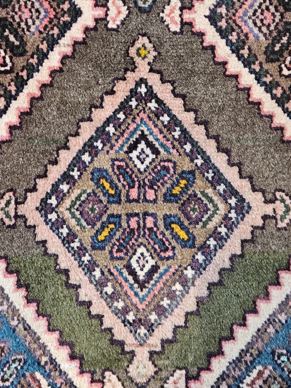 A long hand-woven wool runner rug featuring repeating diamond geometric medallions on an olive green background with decorative navy borders. View III