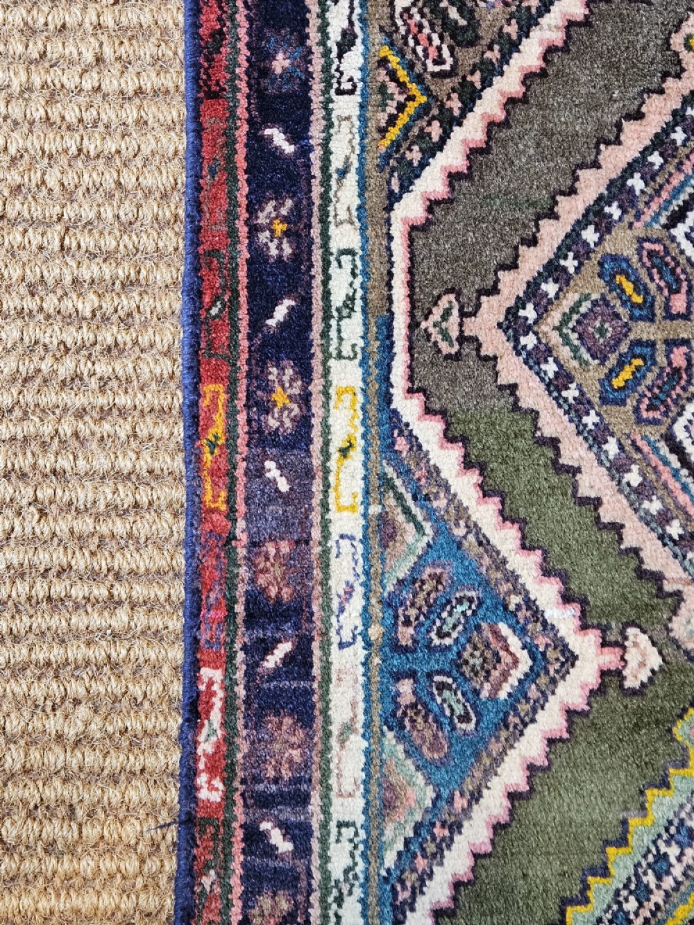 A long hand-woven wool runner rug featuring repeating diamond geometric medallions on an olive green background with decorative navy borders. View V