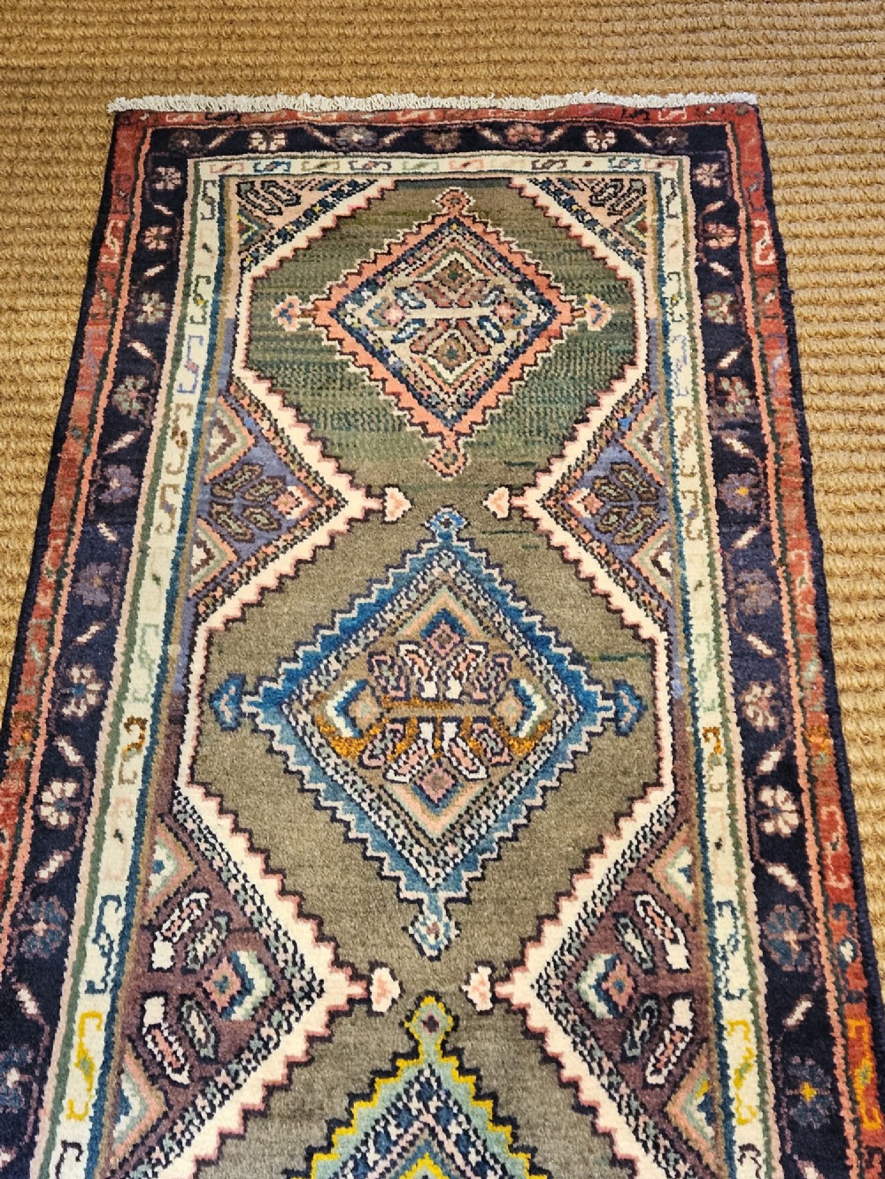 A long hand-woven wool runner rug featuring repeating diamond geometric medallions on an olive green background with decorative navy borders. View VII