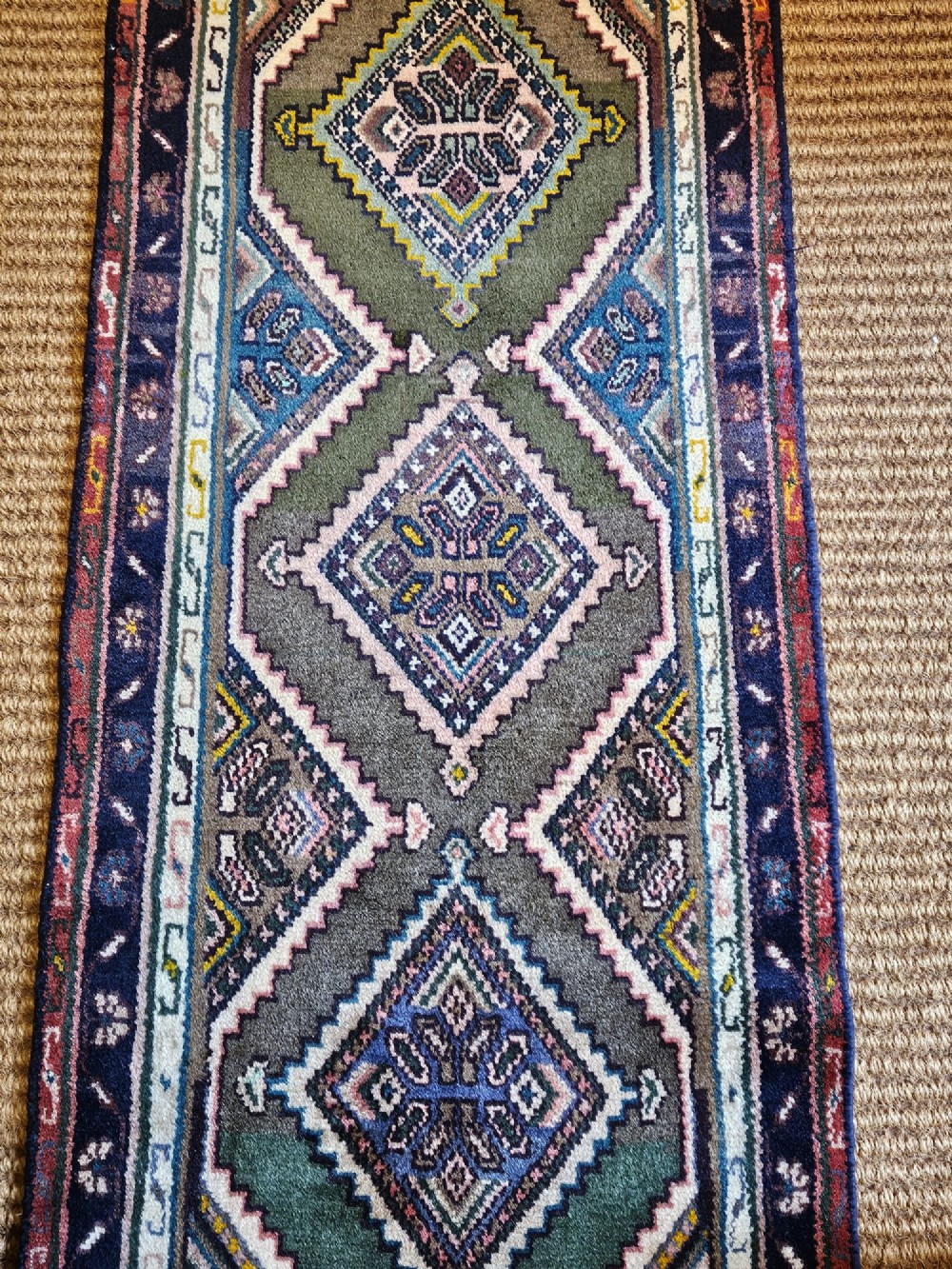 A long hand-woven wool runner rug featuring repeating diamond geometric medallions on an olive green background with decorative navy borders. View VIII