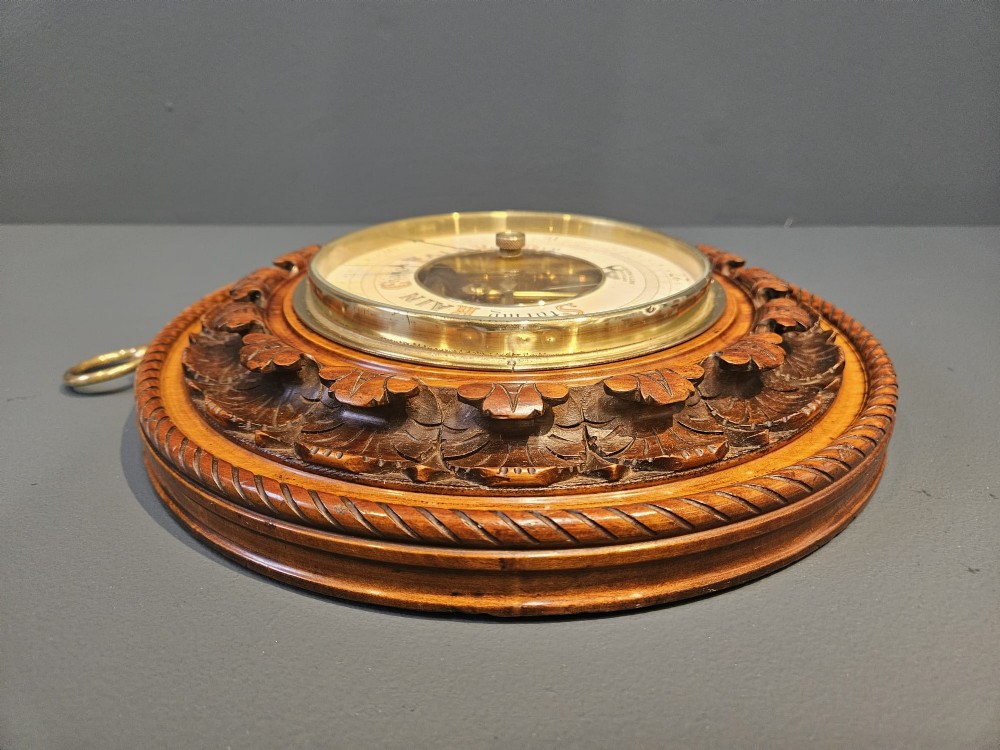 A circular Victorian carved walnut aneroid barometer featuring a rope twist border, leaf motifs, and an open centre dial exposing internal brass mechanisms. View IV